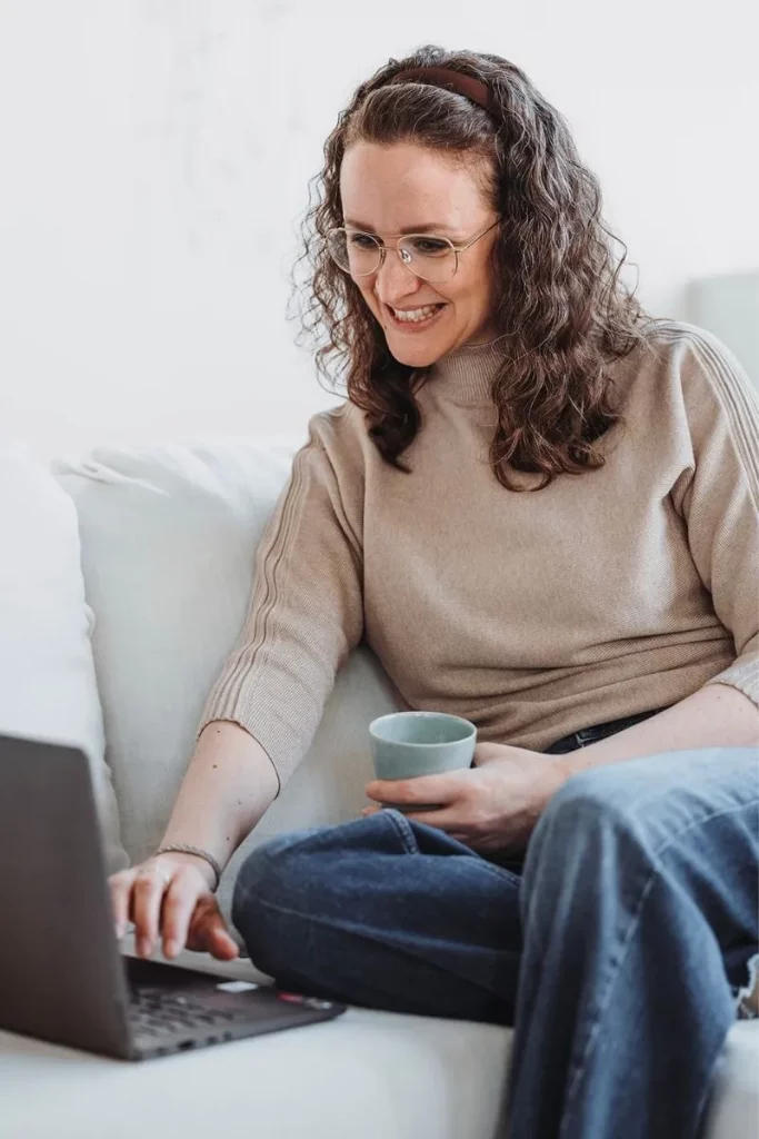 Coach Beatrice Häcki sitzt mit Tasse auf Sofa und arbeitet lächelnd am Laptop in Farbe