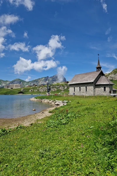 Kleine Steinkapelle am Ufer eines Alpensees mit grünen Wiesen, Bergpanorama und blauem Sommerhimmel